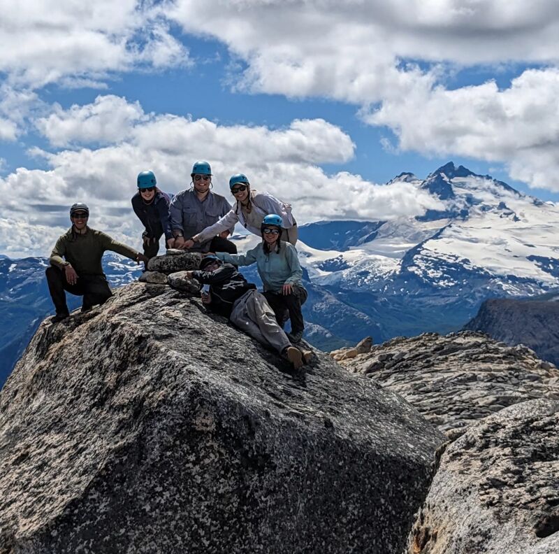 A group of six people are sitting and standing on a rocky mountaintop, posing for a photo. They are all wearing hats or helmets. Behind them, there's a stunning view of snow-capped mountains and a partly cloudy sky. The landscape suggests they are on a hiking or climbing trip, enjoying the scenic vista from their elevated vantage point.